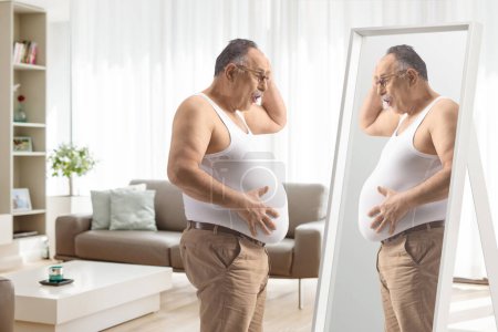Foto de Impresionado hombre maduro mirando su gran vientre en el espejo dentro de una sala de estar - Imagen libre de derechos