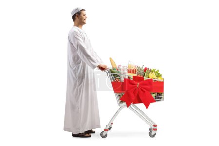 Photo for Full length profile shot of a man in ethnic clothes pushing a shopping trolley with food and red ribbon bow isolated on white background - Royalty Free Image
