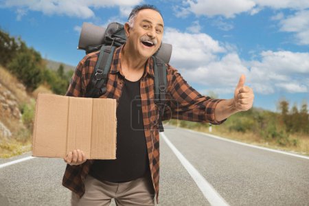 Photo for Smiling mature man with a backpack hitchhiking on the road - Royalty Free Image