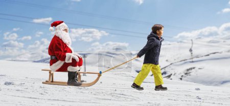 Photo for Boy pulling a sleigh with Santa claus on a snowy mountain hill - Royalty Free Image