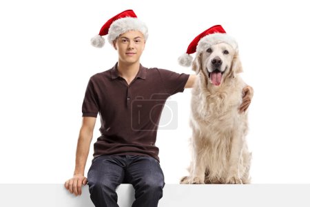 Photo for Teenager and a retriever dog wearing santa claus hats and sitting on a blank panel isolated on white background - Royalty Free Image