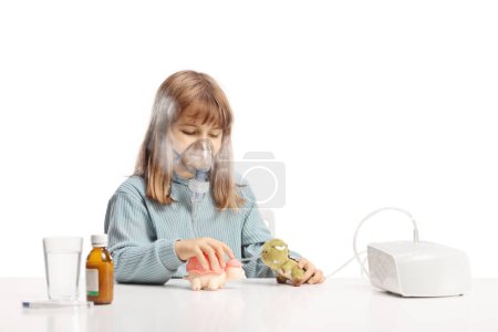 Téléchargez les photos : Petite fille jouant avec des jouets tout en utilisant un nébuliseur avec brouillard de vapeur isolé sur fond blanc - en image libre de droit