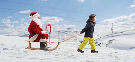 Photo for Boy pulling a sleigh with Santa claus sitting and holding a present on a snowy mountain hill - Royalty Free Image
