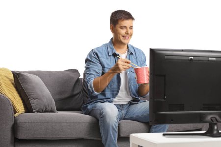 Téléchargez les photos : Jeune homme assis devant la télévision et manger des nouilles avec des baguettes isolées sur fond blanc - en image libre de droit