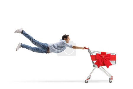 Photo for Full length shot of a casual young man flying and holding an empty shopping cart with a red ribbon bow isolated on white background - Royalty Free Image