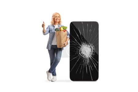Foto de Mujer con una bolsa de comestibles apoyada en un gran teléfono móvil con la pantalla rota y mostrando los pulgares hacia arriba aislado sobre fondo blanco - Imagen libre de derechos