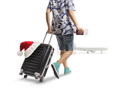 Foto de Vista trasera de un pasajero macho tirando de una maleta con un sombrero de santa y caminando hacia un avión aislado sobre fondo blanco - Imagen libre de derechos