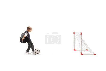Téléchargez les photos : Écolier en uniforme frappant un ballon de football sur mini buts isolés sur fond blanc - en image libre de droit