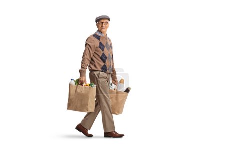 Foto de Largura completa de un pensionista con bolsas de comestibles caminando y mirando a la cámara aislada en el fondo blanco - Imagen libre de derechos