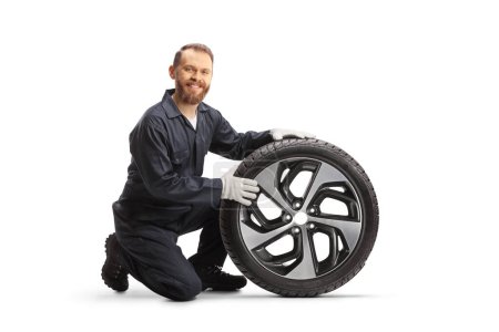 Photo for Car mechanic kneeling next to a tire with a rim and smiling isolated on white background - Royalty Free Image