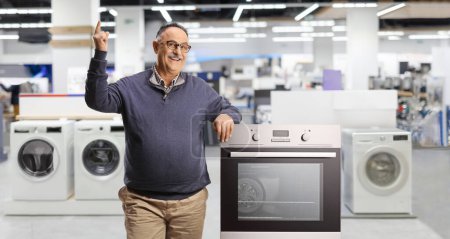 Photo for Smiling mature man leaning on an oven and pointing up inside a shop for home appliances - Royalty Free Image