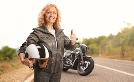 Téléchargez les photos : Femme mûre avec une moto tenant un casque et geste pouces sur la route - en image libre de droit