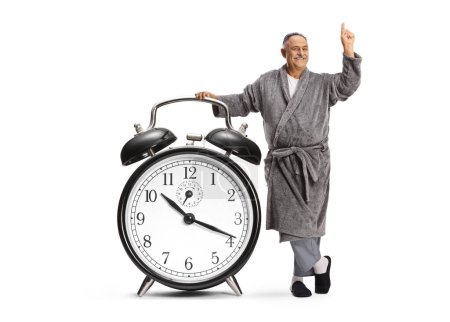 Foto de Hombre maduro en una bata apoyado en un gran despertador y apuntando aislado sobre fondo blanco - Imagen libre de derechos