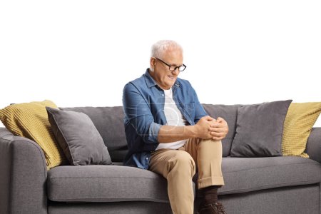 Téléchargez les photos : Homme mûr dans la douleur assis sur un canapé et tenant son genou isolé sur fond blanc - en image libre de droit