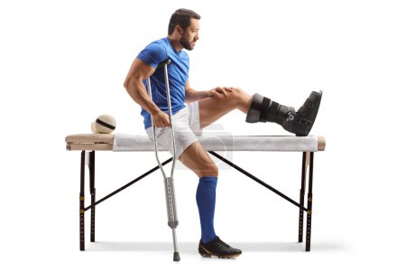 Téléchargez les photos : Athlète masculin avec des béquilles assis sur un lit de thérapie physique isolé sur fond blanc - en image libre de droit
