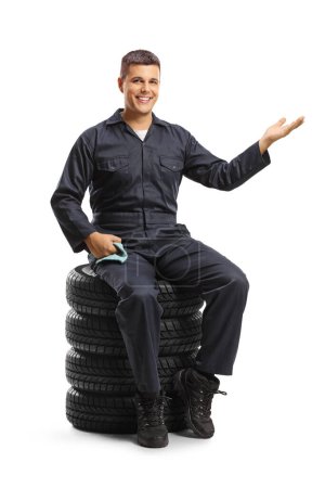 Téléchargez les photos : Travailleur mécanicien automobile assis sur un tas de pneus et geste bienvenue isolé sur fond blanc - en image libre de droit