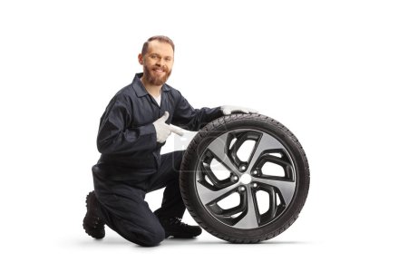 Téléchargez les photos : Mécanicien de voiture agenouillé à côté d'un pneu et pointant isolé sur fond blanc - en image libre de droit