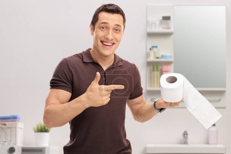 Téléchargez les photos : Joyeux jeune homme pointant un rouleau de papier toilette à l'intérieur d'une salle de bain à la maison - en image libre de droit