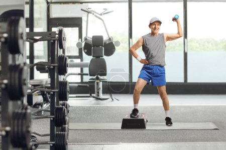 Photo for Full length portrait of an elderly man in sportswear exercising with a step aerobic platform and a small weight at a gym - Royalty Free Image