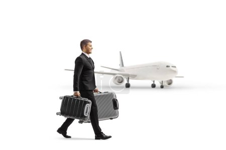 Photo for Full length profile shot of a businessman in a suit walking and carrying suitcases in front of an airplane isolated on white background - Royalty Free Image