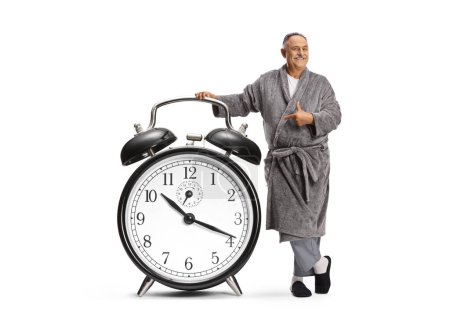 Foto de Hombre maduro en un albornoz apuntando a un gran despertador aislado sobre fondo blanco - Imagen libre de derechos