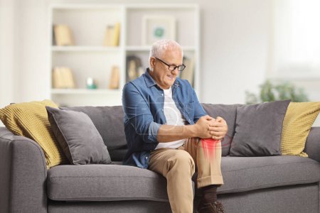 Téléchargez les photos : Homme mûr dans la douleur assis sur un canapé à la maison et tenant son genou enflammé rouge - en image libre de droit