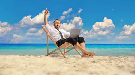 Photo for Cheerful businessman with headphones and laptop sitting on a beach chair by the sea - Royalty Free Image