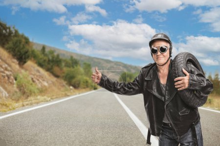Photo for Mature hitchhiker in a black leather holding a flat tire on an open road - Royalty Free Image