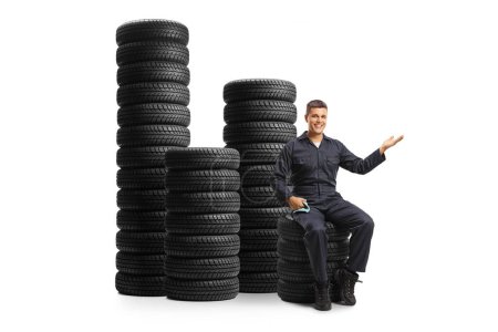 Photo for Auto mechanic sitting on a pile of tires and gesturing welcome isolated on white background - Royalty Free Image