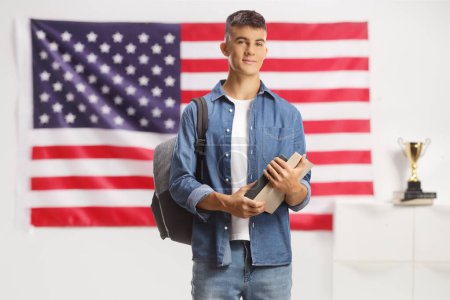 Foto de Estudiante masculino con una mochila de pie y sosteniendo libros frente a una bandera de EE.UU. - Imagen libre de derechos