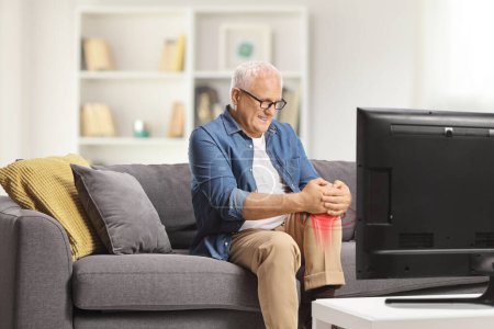 Téléchargez les photos : Homme mûr dans la douleur assis sur un canapé devant la télévision et tenant son genou enflammé rouge - en image libre de droit