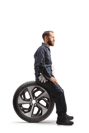 Photo for Full length profile shot of a car mechanic in a uniform sitting on a tire isolated on white background - Royalty Free Image
