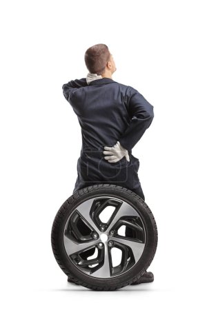 Photo for Rear view of a car mechanic in a uniform sitting on a tire and holding painful back isolated on white background - Royalty Free Image