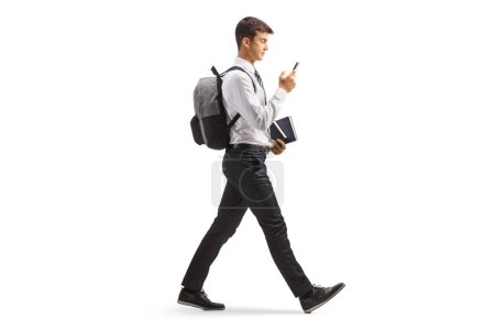 Téléchargez les photos : Photo de profil pleine longueur d'un étudiant masculin en uniforme de collège marchant avec un smartphone isolé sur fond blanc - en image libre de droit
