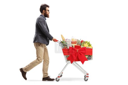 Foto de Foto de perfil completo de un tipo barbudo empujando un carrito de compras de Navidad con productos alimenticios aislados sobre fondo blanco - Imagen libre de derechos