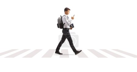 Téléchargez les photos : Étudiant dans un uniforme universitaire marchant avec un smartphone à un passage à niveau isolé sur fond blanc - en image libre de droit