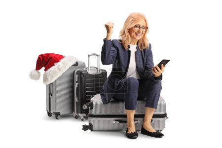 Photo for Happy woman sitting on a suitcase with a santa hat and looking at a smartphone isolated on white background - Royalty Free Image