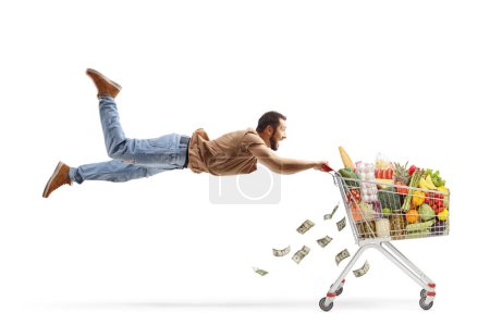 Photo for Casual man flying and holding onto a shopping cart with food and money isolated on white background - Royalty Free Image