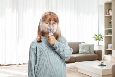 Photo for Little girl using an inhaler mask with vapor mist at home in a living room - Royalty Free Image