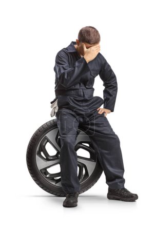 Téléchargez les photos : Mécanicien de voiture en uniforme assis sur un pneu et tenant sa tête isolée sur fond blanc - en image libre de droit