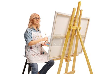 Téléchargez les photos : Femme d'âge moyen assise sur une chaise et peignant sur une toile isolée sur fond blanc - en image libre de droit