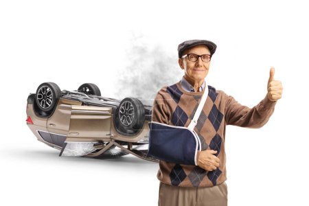 Téléchargez les photos : Homme âgé avec un bras cassé portant une attelle et les pouces gestuels devant une voiture endommagée par un accident isolé sur fond blanc - en image libre de droit