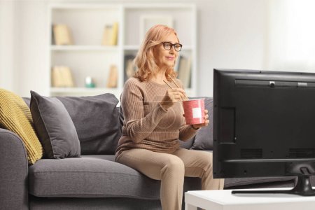 Photo for Woman sitting on a couch in front of tv and eating noodles - Royalty Free Image