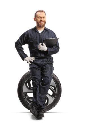 Téléchargez les photos : Mécanicien de voiture souriant et assis sur un pneu avec un document dans les mains isolé sur fond blanc - en image libre de droit