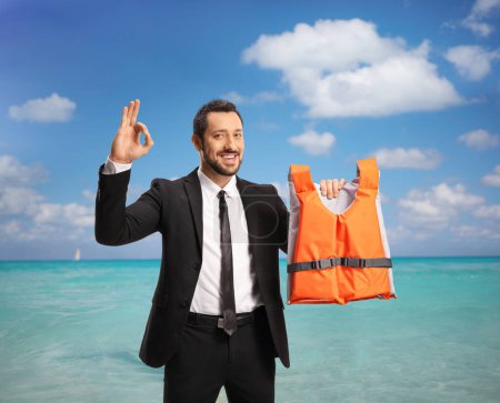 Foto de Hombre con traje y corbata sosteniendo un chaleco salvavidas junto al mar y haciendo un gesto de una señal aceptable - Imagen libre de derechos