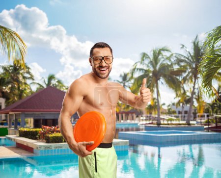 Téléchargez les photos : Joyeux jeune homme en maillot de bain tenant un disque volant en plastique et gesticulant les pouces vers le haut près de la piscine - en image libre de droit