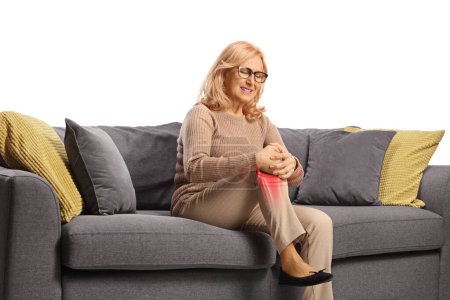 Foto de Mujer de mediana edad con dolor sosteniendo la rodilla y sentada en un sofá aislado sobre fondo blanco - Imagen libre de derechos
