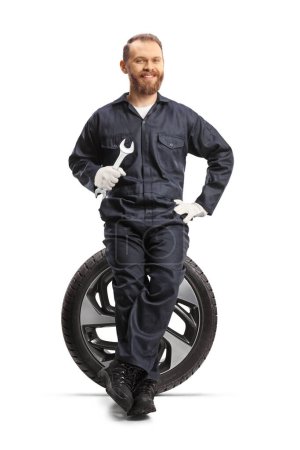 Téléchargez les photos : Mécanicien de voiture tenant une clé et assis sur un pneu isolé sur fond blanc - en image libre de droit