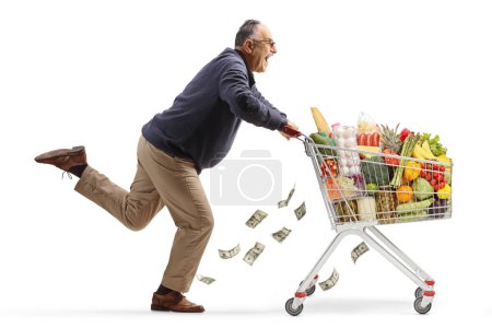 Foto de Emocionado hombre maduro corriendo con un carrito de compras, mientras que el dinero están cayendo del carro aislado sobre fondo blanco - Imagen libre de derechos