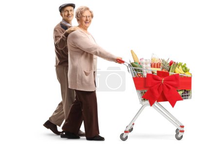 Foto de Foto completa de una pareja de ancianos caminando y empujando un carrito de compras con comida y cinta roja aislada sobre fondo blanco - Imagen libre de derechos